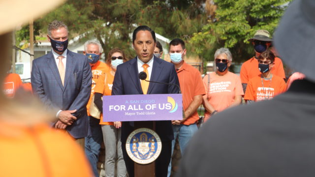 Mayor Todd Gloria speaks at the podium at an event. Behind him stands a group of people, including a Moms Demand Action volunteer.