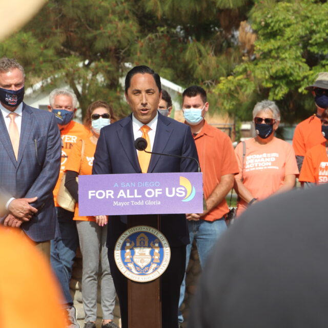 Mayor Todd Gloria speaks at the podium at an event. Behind him stands a group of people, including a Moms Demand Action volunteer.